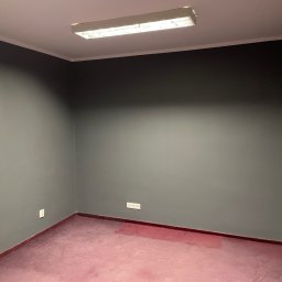 Łukasz Berliński - Puste pomieszczenie biurowe po remoncie, ściany pomalowane na szaro, z widoczną instalacją elektryczną i fragmentem podłogi w odcieniach różu.