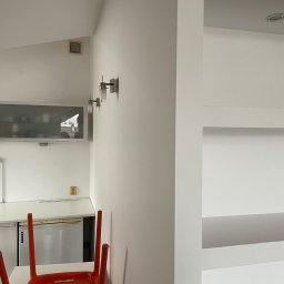 Łukasz Berliński - Białe wnętrze kuchni po remoncie, widoczne gładkie ściany, oświetlenie, zabudowa kuchenna i pomarańczowe krzesło, Praga Południe, Warszawa.