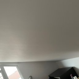 Łukasz Berliński - Gładka, jasna powierzchnia sufitu po szpachlowaniu i malowaniu, widok z dołu, fragment okna w lewym dolnym rogu, częściowo widoczne meble w tle.
