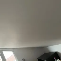 Gładka, jasna powierzchnia sufitu po szpachlowaniu i malowaniu, widok z dołu, fragment okna w lewym dolnym rogu, częściowo widoczne meble w tle.