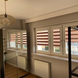 Łukasz Berliński - Pokój z trzema oknami z roletami poziomymi, grzejnikami pod oknami, lustrzaną szafą i lampą wiszącą z ażurowym kloszem.