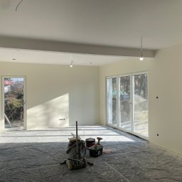 Łukasz Berliński - Wnętrze w trakcie prac wykończeniowych: świeżo pomalowane ściany w odcieniach bieli, podłoga zabezpieczona folią malarską, widoczny odkurzacz przemysłowy, pędzle i wiaderka z farbą.