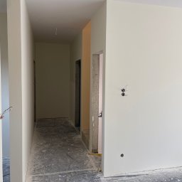 Łukasz Berliński - Świeżo pomalowany korytarz w odcieniach bieli, widoczne wystające kable elektryczne i surowy beton wokół ościeżnicy, podłoga zabezpieczona folią malarską.