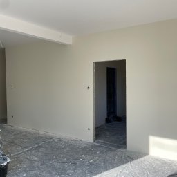 Łukasz Berliński - Wnętrze po świeżym pomalowaniu ścian na jasny odcień szarości, widoczny otwór drzwiowy, zabezpieczona podłoga folią malarską, odkurzacz przemysłowy i wiadro z farbą w rogu pomieszczenia.