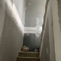 Łukasz Berliński - Klatka schodowa w trakcie prac wykończeniowych: ściany częściowo pokryte gładzią, widoczne niedoskonałości i ślady po szpachlowaniu, na schodach puszka farby i karton z elementami wentylacyjnymi.