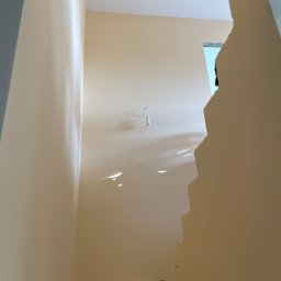 Łukasz Berliński - Świeżo pomalowana klatka schodowa w odcieniach beżu, z widocznymi niedoskonałościami ściany i prowizorycznym oświetleniem, ukazująca etap prac wykończeniowych w Brwinowie.