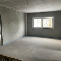 Łukasz Berliński - Surowe wnętrze w Brwinowie przed malowaniem, z widocznymi surowymi ścianami, oknem z widokiem na drzewa i niebieską taśmą zabezpieczającą podłogę.