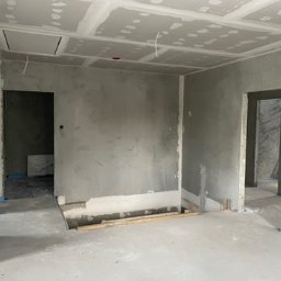 Łukasz Berliński - Surowe wnętrze w trakcie prac przygotowawczych: ściany i sufit pokryte wstępną warstwą szpachli, widoczne łączenia płyt gipsowo-kartonowych, zabezpieczona podłoga, otwory drzwiowe bez ościeżnic.