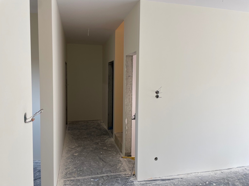 Świeżo pomalowany korytarz w odcieniach bieli, widoczne wystające kable elektryczne i surowy beton wokół ościeżnicy, podłoga zabezpieczona folią malarską.