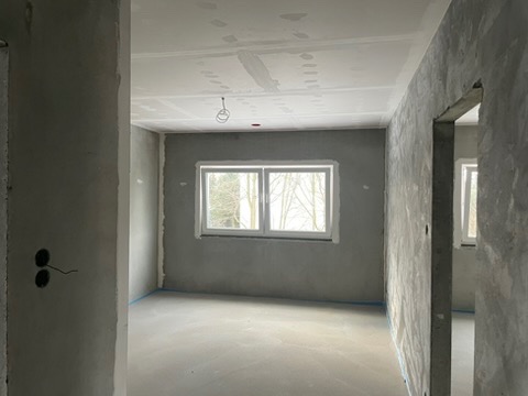 Pokój po szpachlowaniu ścian i sufitu, przygotowany do dalszych prac wykończeniowych, widoczne okno z widokiem na drzewa, podłoga zabezpieczona folią malarską, surowy stan wykończenia.