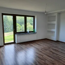 Pusty pokój po remoncie z brązowymi panelami podłogowymi ułożonymi w jodełkę, białymi ścianami z przygotowanymi gniazdkami i wnękami, widokiem na zieleń przez okno.