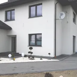 Dwukondygnacyjny dom z jasną elewacją, ciemnymi oknami i dachem, z satelitą na ścianie, otoczony ozdobnym ogrodem z białym żwirem i kulistymi lampami.