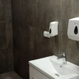 Umywalka z szafką w nowoczesnej łazience, ściany wyłożone ciemnymi, imitującymi kamień płytkami, widoczne dozowniki mydła i papieru.