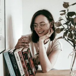 Uśmiechnięta kobieta w okularach siedzi przy drewnianym stole z książkami, trzymając szklankę z napojem, z zegarem i rośliną w tle.