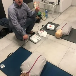 Instruktor prezentuje elektrody do defibrylatora podczas szkolenia z resuscytacji krążeniowo-oddechowej, manekiny na matach, AED na stole w tle.