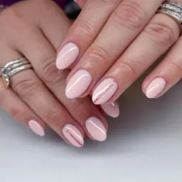 Delikatny, pudrowo-różowy manicure hybrydowy z akcentem w postaci cienkiego, brokatowego paska na dwóch paznokciach, ozdobne pierścionki na palcach.