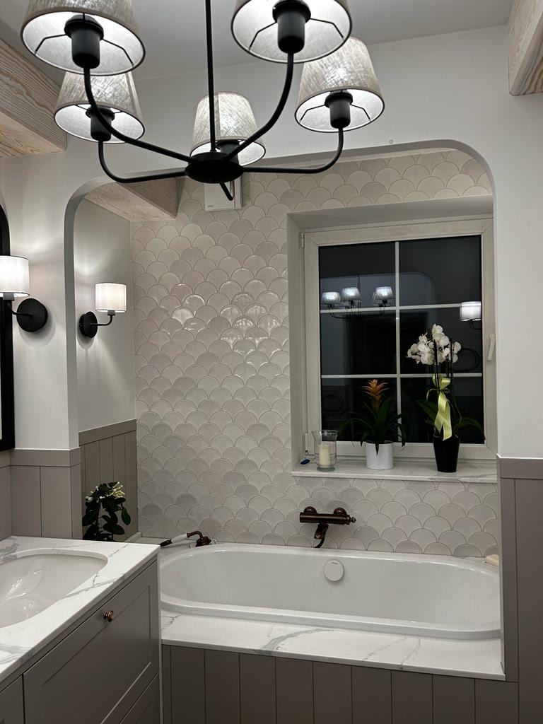 Elegancka łazienka z wanną obudowaną marmurem, ściany wyłożone płytkami w kształcie rybiej łuski, oświetlenie sufitowe w stylu retro, okno z roślinami i świecą.