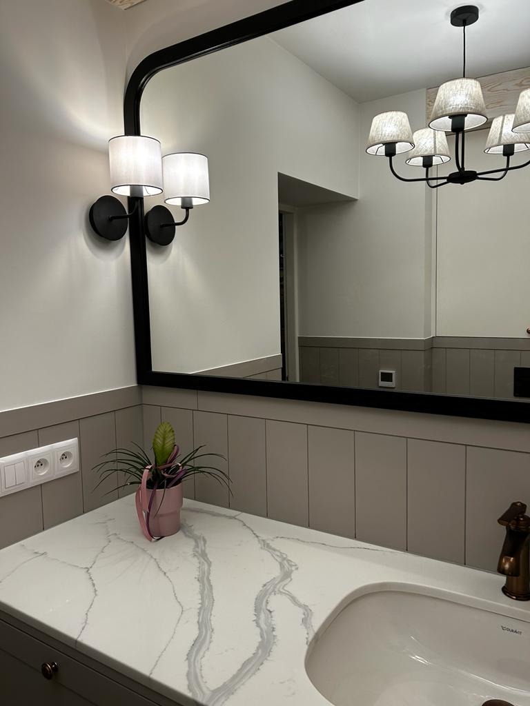 Nowoczesna łazienka z marmurowym blatem, czarną ramą lustra z oświetleniem ściennym i designerską lampą sufitową, uzupełniona dekoracyjną rośliną w różowej doniczce.