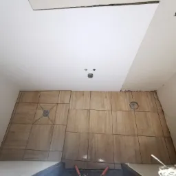 Układanie brązowych płytek podłogowych w łazience, widoczny odpływ liniowy i otwór wentylacyjny w podłodze, biały sufit z punktowym oświetleniem w trakcie montażu.