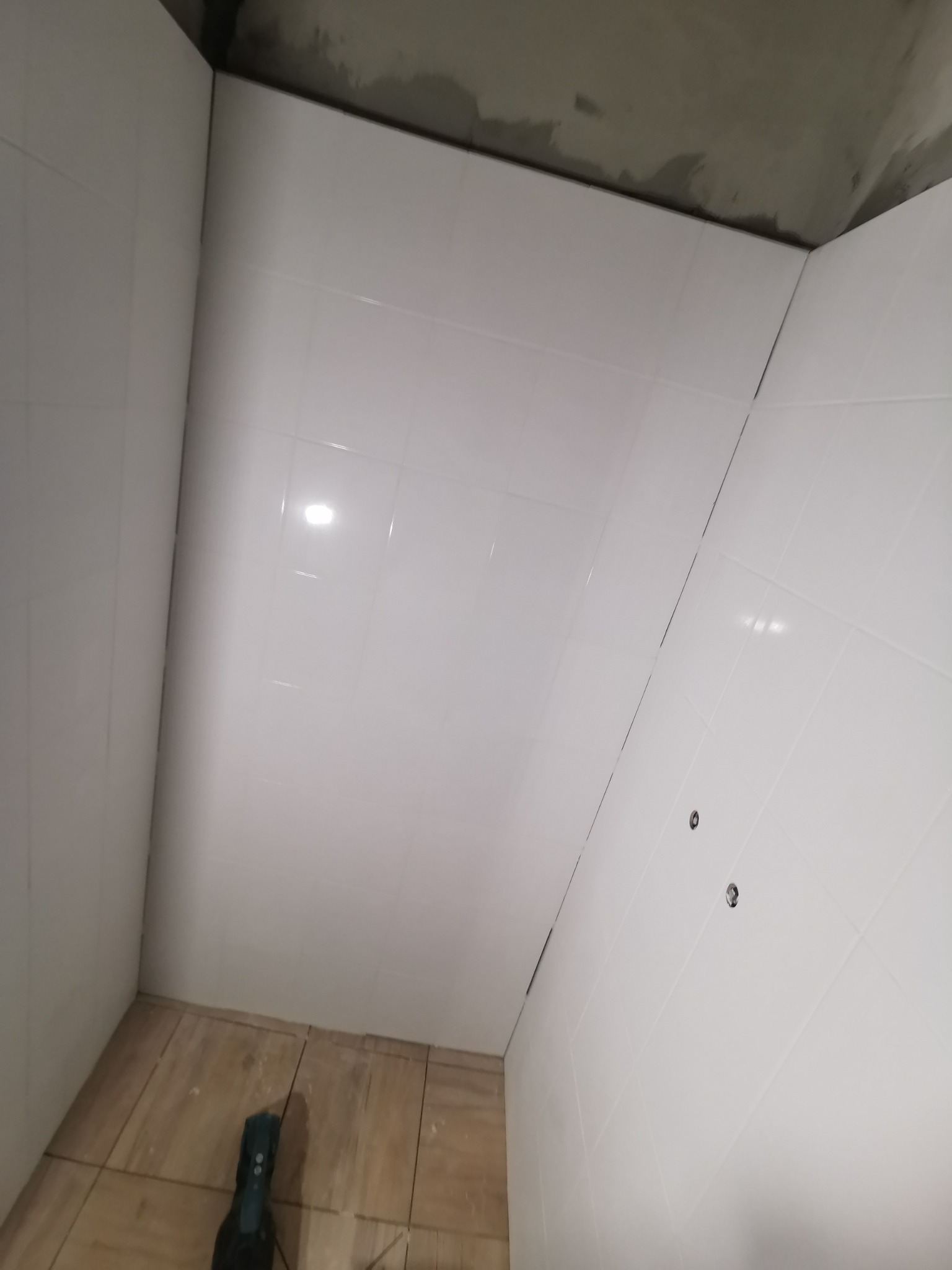 Wnętrze łazienki w trakcie prac wykończeniowych, widoczne białe płytki na ścianach z ciemną fugą i otwory na instalacje, sufit w stanie surowym, na podłodze płytki imitujące drewno, w rogu widoczny...