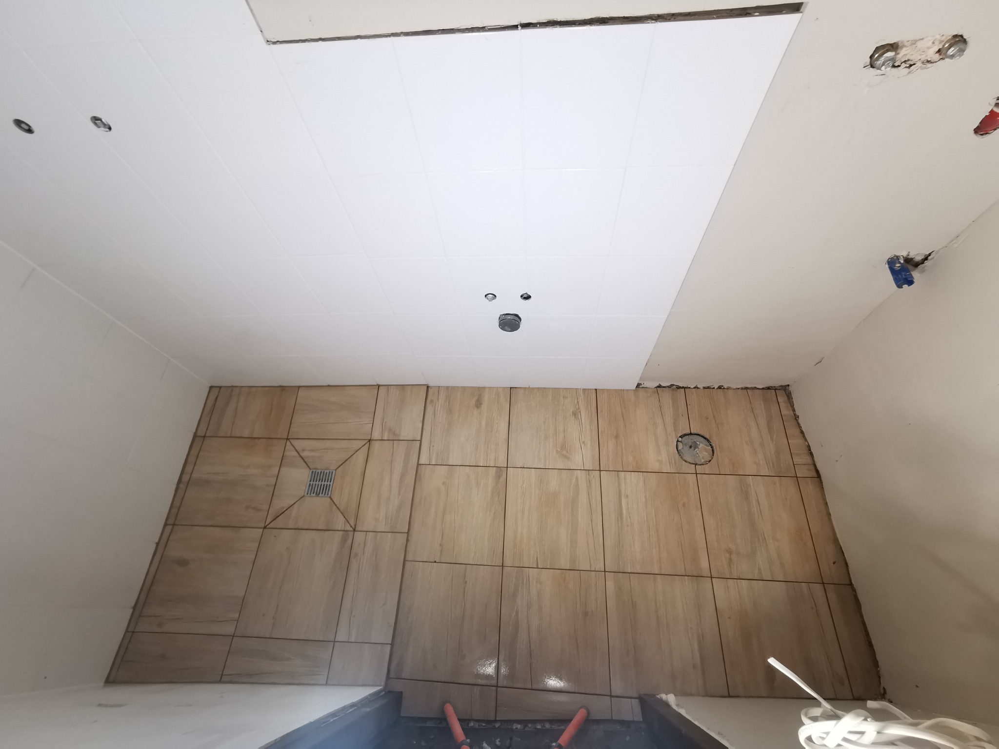 Układanie brązowych płytek podłogowych w łazience, widoczny odpływ liniowy i otwór wentylacyjny w podłodze, biały sufit z punktowym oświetleniem w trakcie montażu.