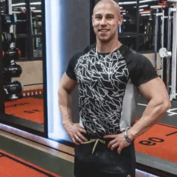 Uśmiechnięty mężczyzna w sportowej koszulce z geometrycznym wzorem, stojący w siłowni na tle luster i sprzętu do ćwiczeń.