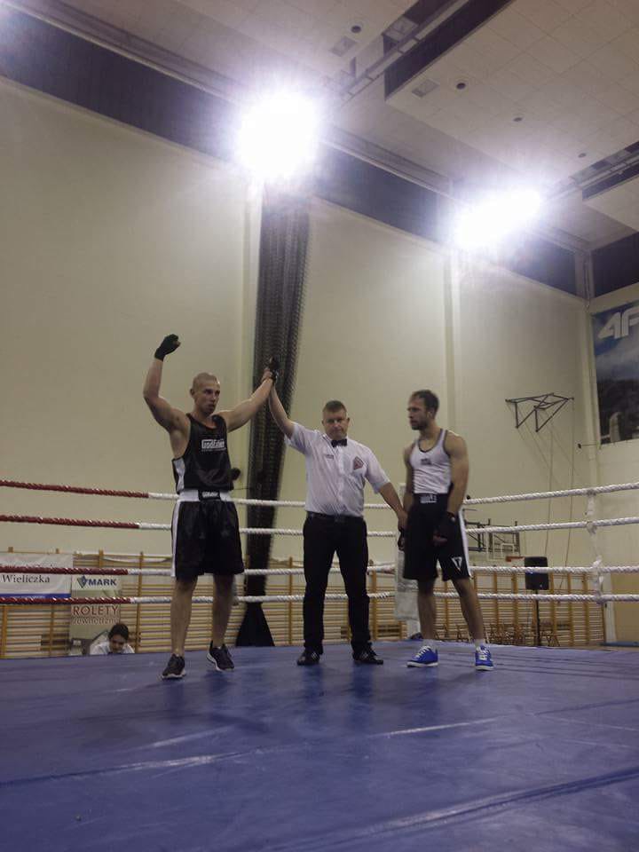 Bokser z podniesioną ręką ogłoszony zwycięzcą walki na ringu, sędzia w białej koszuli i czarnej muszce, drugi zawodnik stoi obok.