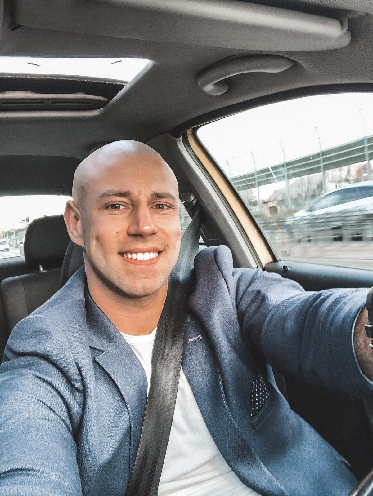 Uśmiechnięty mężczyzna w samochodzie, ubrany w marynarkę i białą koszulkę, zapięty pasem bezpieczeństwa, widoczny fragment deski rozdzielczej i otwierany dach.