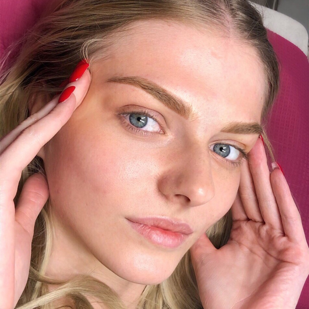 Portret kobiety z delikatnym makijażem permanentnym brwi, podkreślone spojrzenie, czerwone paznokcie, blada cera.