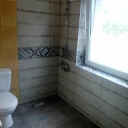 Wnętrze łazienki po remoncie: biała toaleta, kabina prysznicowa z chromowaną armaturą i deszczownicą, ściany wyłożone płytkami imitującymi drewno z dekoracyjnym pasem płytek z motywami...