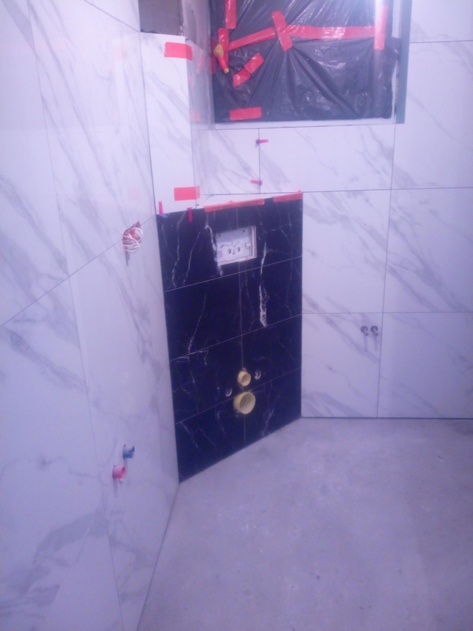 Wnętrze łazienki w trakcie prac wykończeniowych, widoczne białe płytki imitujące marmur na ścianach, czarna obudowa stelaża WC oraz zakryte folią okno.