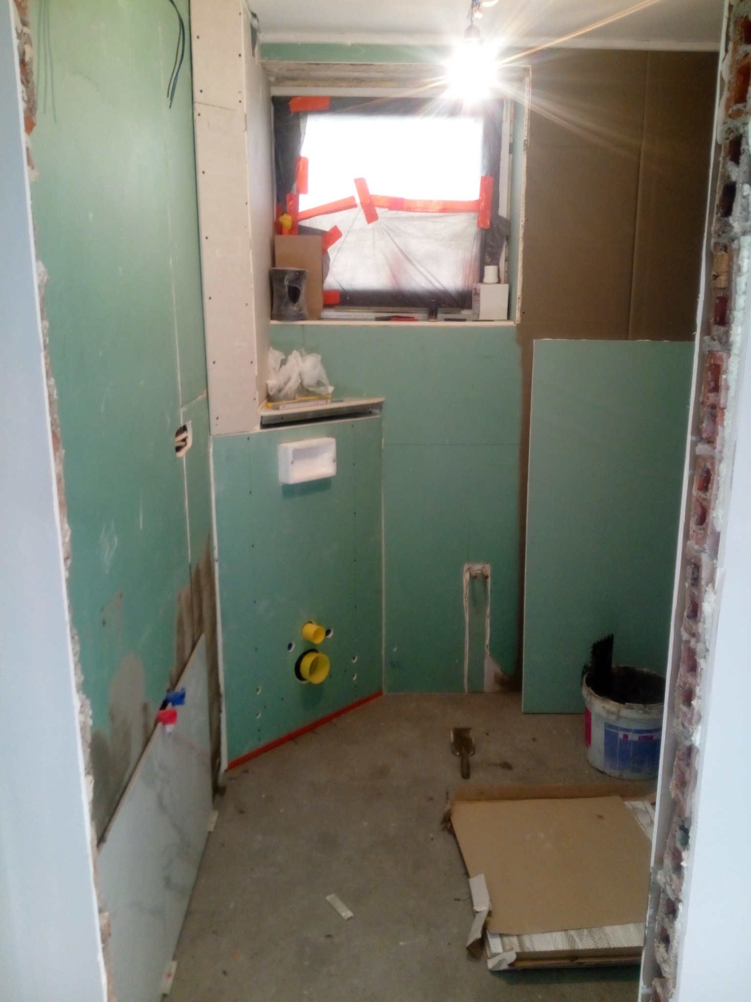 Wnętrze łazienki w trakcie remontu: widoczne zielone płyty kartonowo-gipsowe na ścianach, przygotowane pod instalację sanitarną, okno zabezpieczone folią i taśmą, oraz narzędzia i wiadro na podłodze.