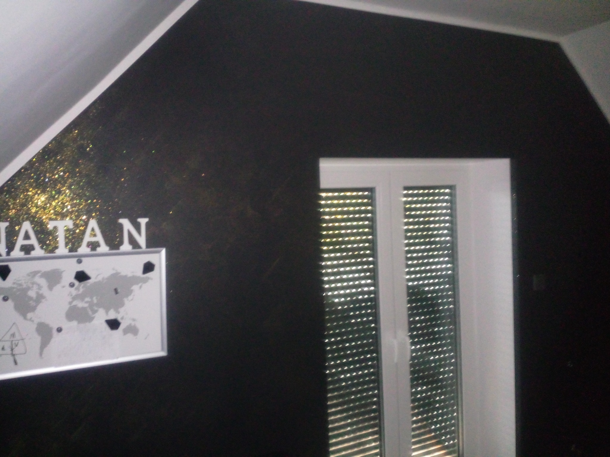 Ciemna ściana w pokoju na poddaszu z oknem z żaluzjami, dekorowana złotym brokatem i mapą świata z zaznaczonymi punktami oraz napisem 'NATAN' powyżej.