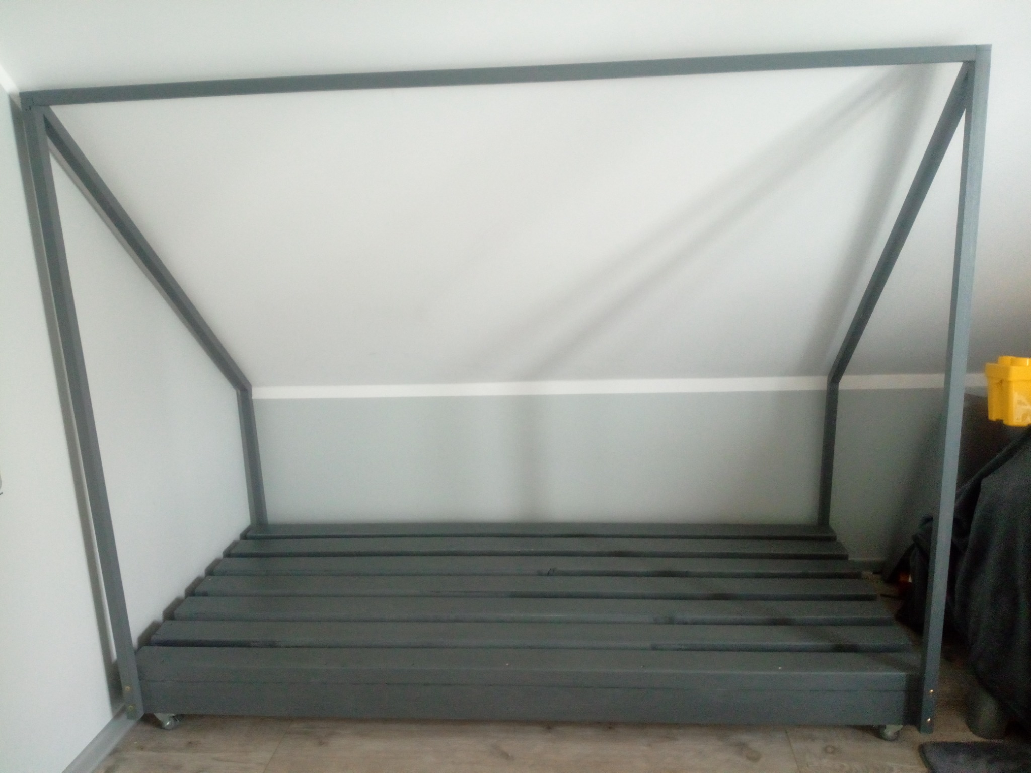 Szara, metalowa konstrukcja łóżka z drewnianymi listwami na kółkach, ustawiona w pomieszczeniu ze skośnym sufitem, pomalowanym w dwóch odcieniach szarości.