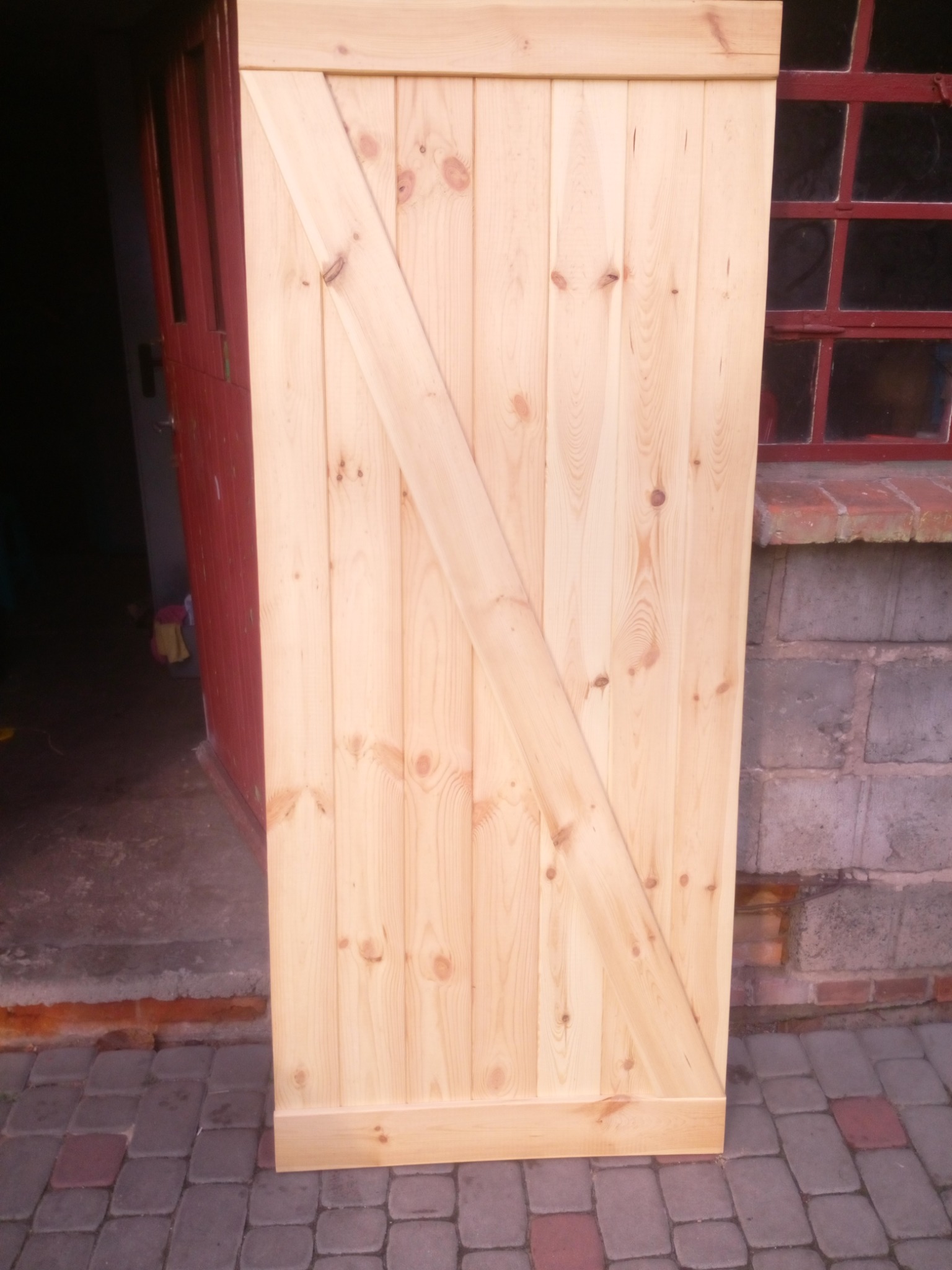 Surowa, drewniana płyta z ukośnym wzmocnieniem, prawdopodobnie drzwi przesuwne typu stodoła, oparta o ścianę z cegieł i okno z czerwonymi ramami, na tle brukowanej powierzchni.