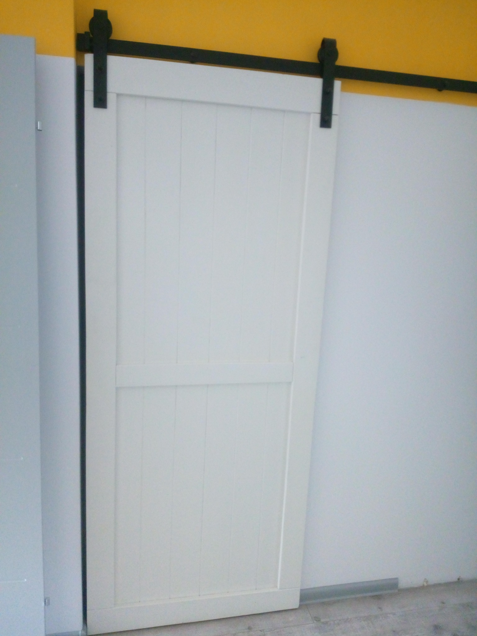 Białe, przesuwne drzwi drewniane zamontowane na czarnej szynie, widoczne na tle białej ściany i fragmentu żółtej ściany u góry.