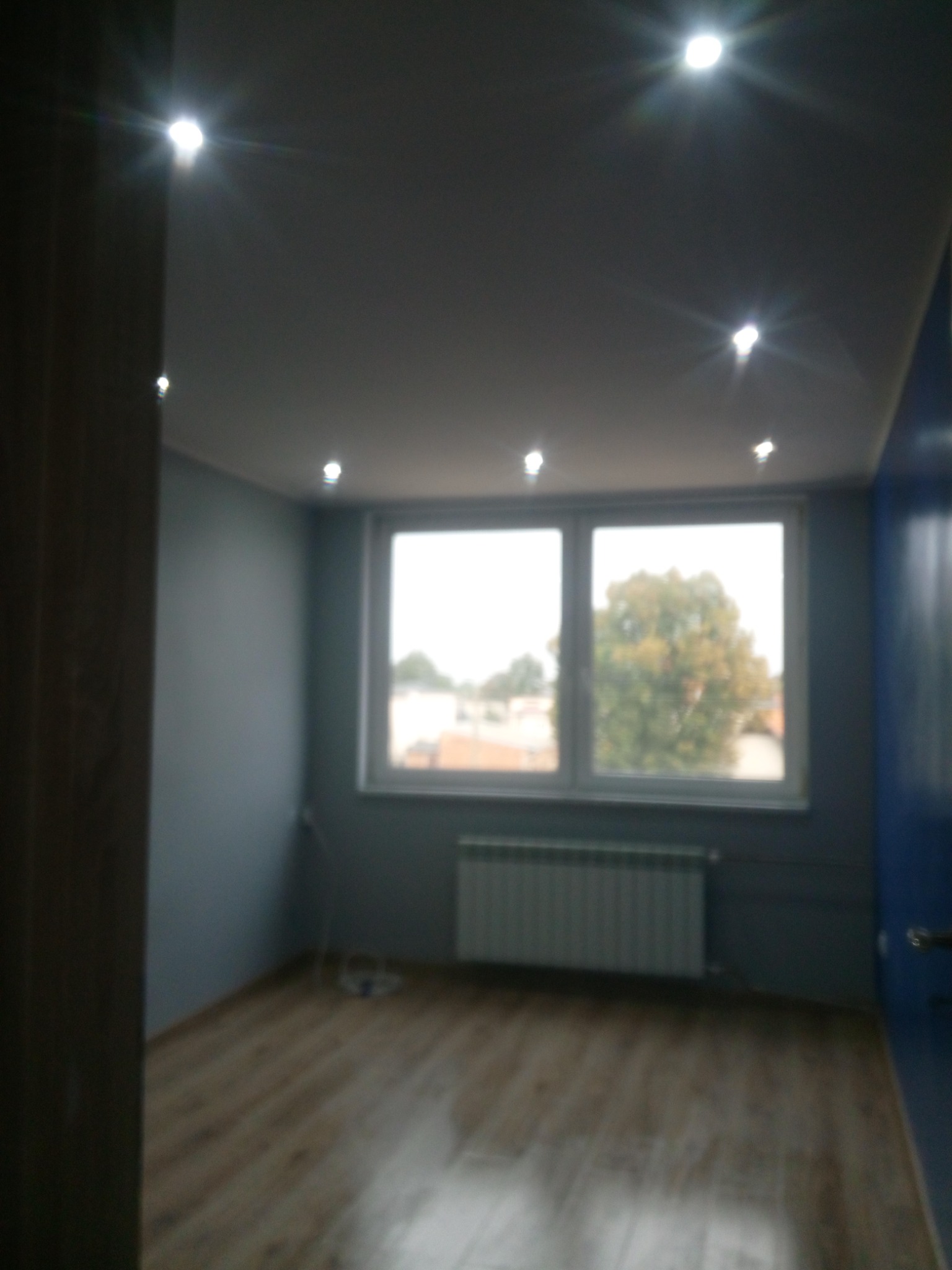 Pokój po odświeżeniu ścian w odcieniach szarości i błękitu, z drewnianą podłogą i oknem z widokiem na drzewo, oświetlony punktowymi lampami sufitowymi.