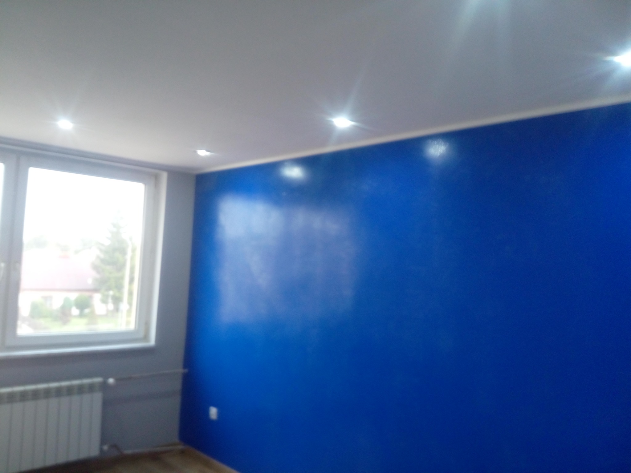 Świeżo pomalowany pokój z intensywnie niebieską ścianą, widoczne punktowe oświetlenie sufitowe i okno z widokiem na zieleń. Grzejnik pod oknem w kolorze szarym.
