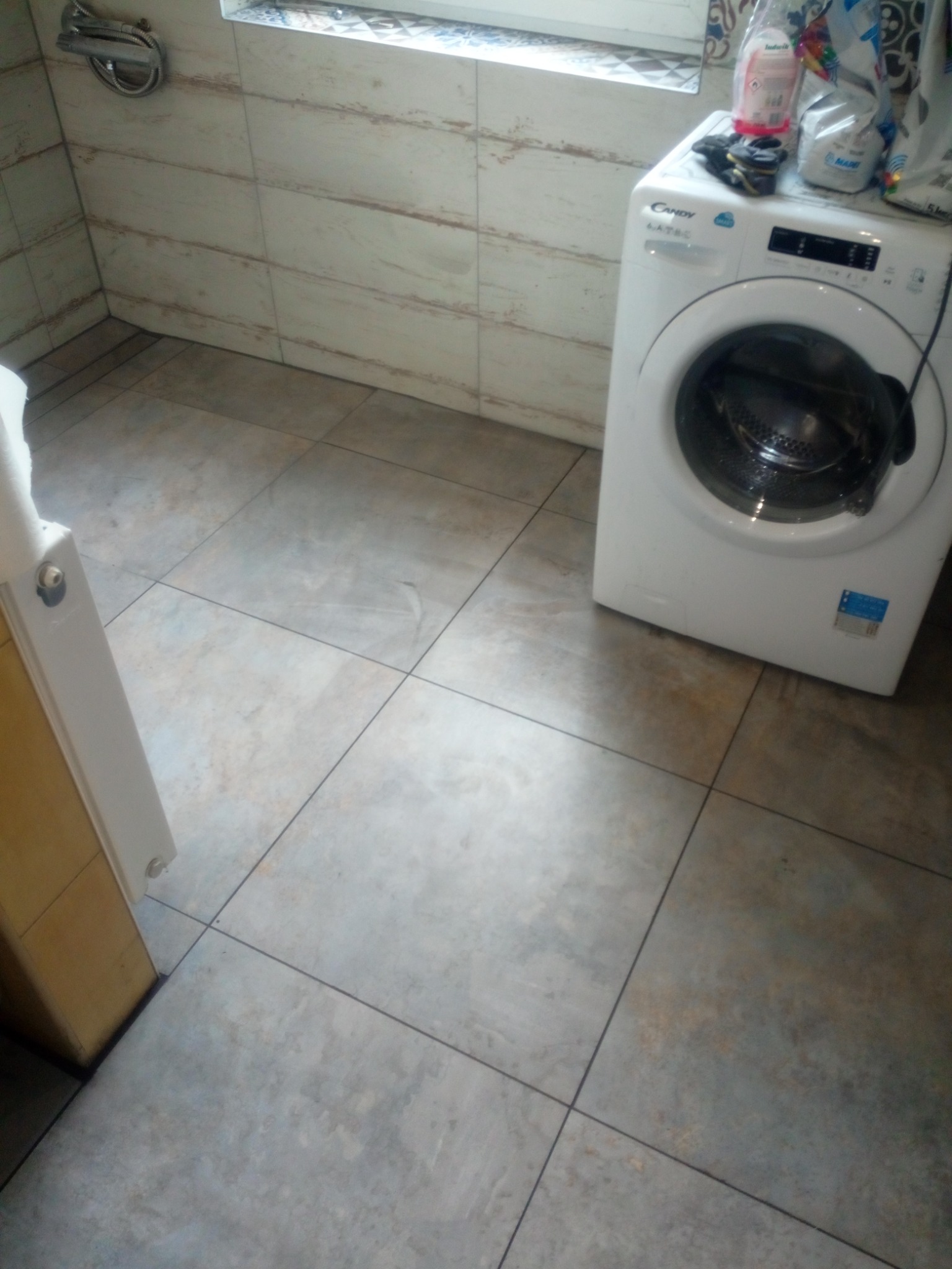 Wyłożona płytkami łazienka z pralką Candy i półką na kosmetyki, płytki imitujące drewno na ścianach i szare kwadratowe płytki na podłodze.