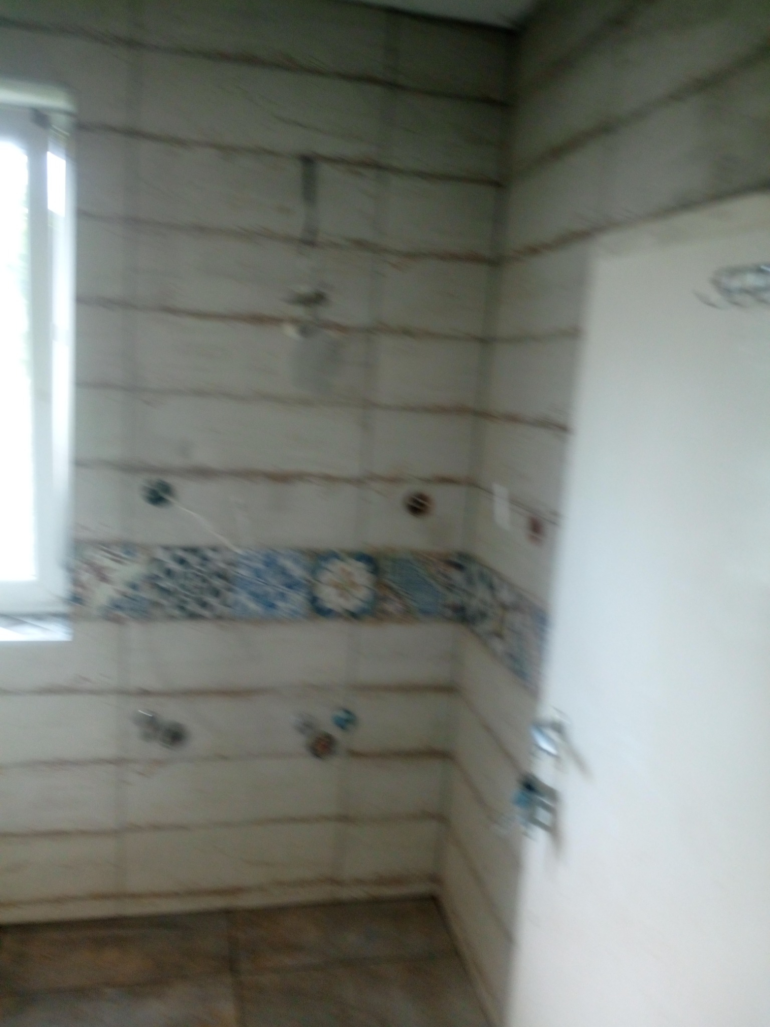 Niedokończona łazienka z ułożonymi płytkami imitującymi drewno i dekoracyjnym pasem płytek z motywem kwiatowym, widoczne wystające kable i rury, drzwi z klamką po prawej stronie.