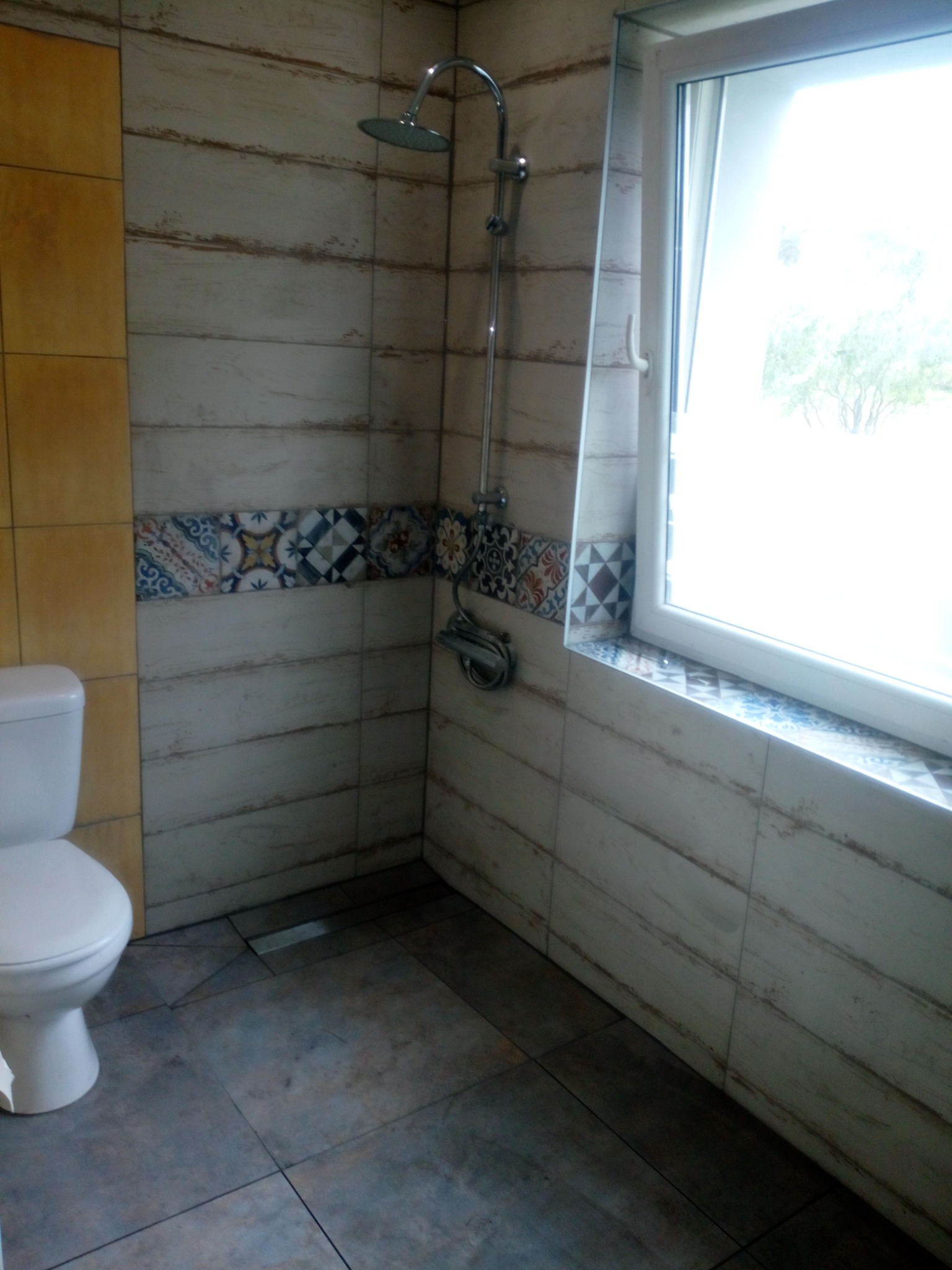 Wnętrze łazienki po remoncie: biała toaleta, kabina prysznicowa z chromowaną armaturą i deszczownicą, ściany wyłożone płytkami imitującymi drewno z dekoracyjnym pasem płytek z motywami...