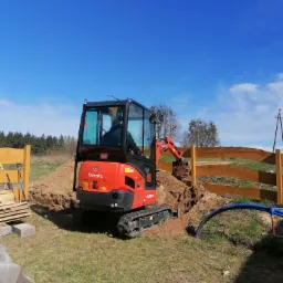 Czerwona mini koparka Kubota wykonuje wykop w ziemi obok drewnianego ogrodzenia na tle błękitnego nieba.