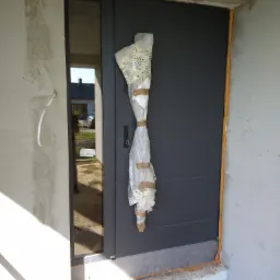 Nowe, grafitowe drzwi wejściowe zabezpieczone folią i taśmą, widoczne pianka montażowa wokół ościeżnicy, surowy stan wykończenia ściany.