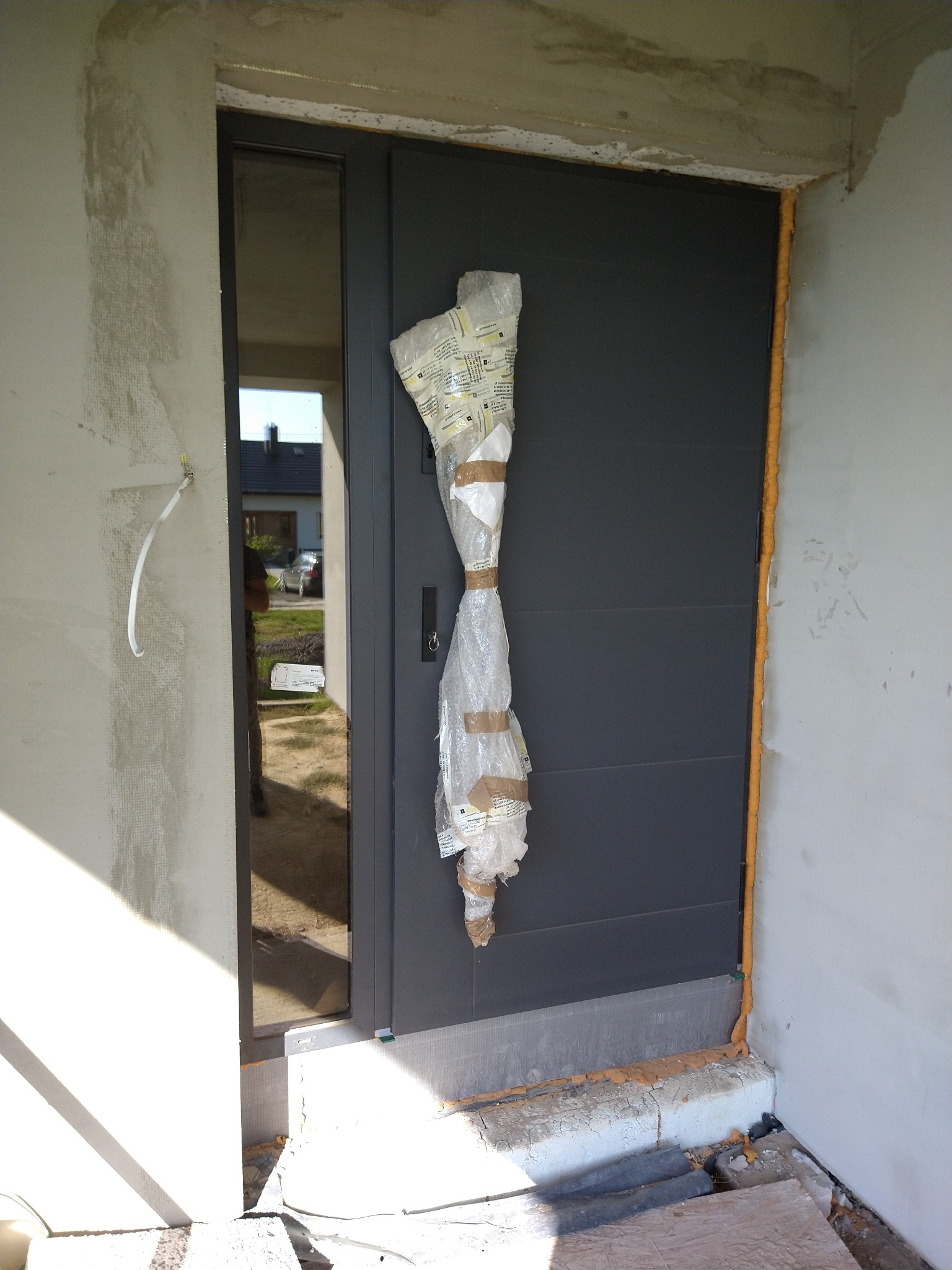 Nowe, grafitowe drzwi wejściowe zabezpieczone folią i taśmą, widoczne pianka montażowa wokół ościeżnicy, surowy stan wykończenia ściany.