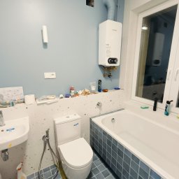 PAUL & ZBIG USŁUGI REMONTOWO - BUDOWLANE PAWEŁ FIJAŁKOWSKI & ZBIGNIEW ZALEWSKI S.C. - Łazienka w trakcie remontu: widoczny biały bojler nad wanną, umywalka, toaleta, niebieskie płytki na podłodze i częściowo na ścianach, ślady po pracach na ścianach, otwory w ścianie, rury, bałagan...