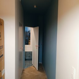 PAUL & ZBIG USŁUGI REMONTOWO - BUDOWLANE PAWEŁ FIJAŁKOWSKI & ZBIGNIEW ZALEWSKI S.C. - Korytarz po remoncie z parkietem w jodełkę, pomalowanym sufitem na ciemny kolor i otwartymi drzwiami do łazienki z szarą glazurą.