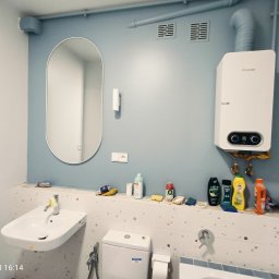 PAUL & ZBIG USŁUGI REMONTOWO - BUDOWLANE PAWEŁ FIJAŁKOWSKI & ZBIGNIEW ZALEWSKI S.C. - Niebieska łazienka z owalnym lustrem, białym zlewem, toaletą i bojlerem Junkers. Na półce nad toaletą stoją środki czystości i inne akcesoria łazienkowe. Widoczne rury i kratki wentylacyjne.