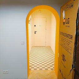 PAUL & ZBIG USŁUGI REMONTOWO - BUDOWLANE PAWEŁ FIJAŁKOWSKI & ZBIGNIEW ZALEWSKI S.C. - Korytarz z żółtym łukowym przejściem, podłoga w szachownicę, fragment podłogi wyłożony parkietem w jodełkę i karton po lodówce LG.