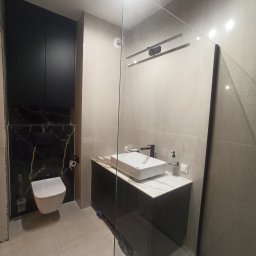 PAUL & ZBIG USŁUGI REMONTOWO - BUDOWLANE PAWEŁ FIJAŁKOWSKI & ZBIGNIEW ZALEWSKI S.C. - Nowoczesna łazienka z podwieszaną toaletą na czarnej marmurowej ścianie, prostokątną umywalką na szafce z blatem imitującym marmur i szklaną kabiną prysznicową.