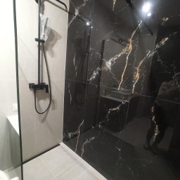 PAUL & ZBIG USŁUGI REMONTOWO - BUDOWLANE PAWEŁ FIJAŁKOWSKI & ZBIGNIEW ZALEWSKI S.C. - Nowoczesna kabina prysznicowa z czarnymi płytkami imitującymi marmur ze złotymi żyłami i czarną armaturą, widoczny fragment białej ściany i brodzika.